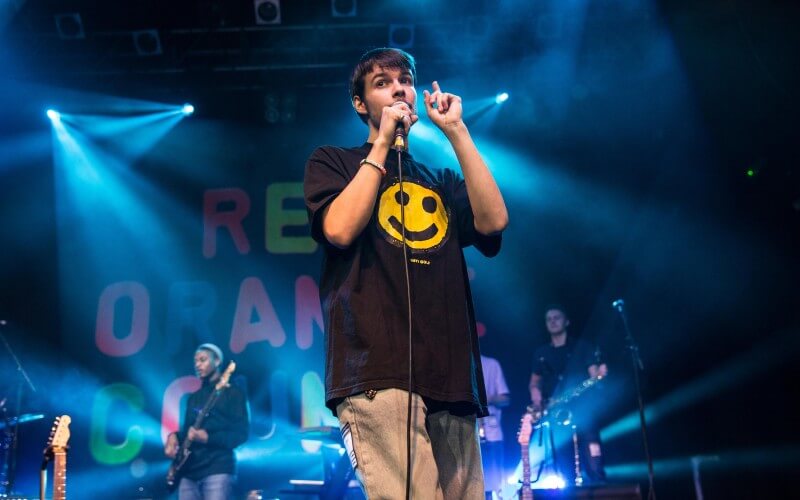 Rex Orange County mengenakan atasan hitam bergambar emoji smile sedang di atas panggung dalam salah satu penampilan konsernya.